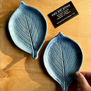 May include: Two blue ceramic leaf-shaped dishes. The dishes have a raised vein pattern and are glazed in a light blue color. The dishes are on a wooden surface. The business card for "angry goat pottery" is visible in the background.