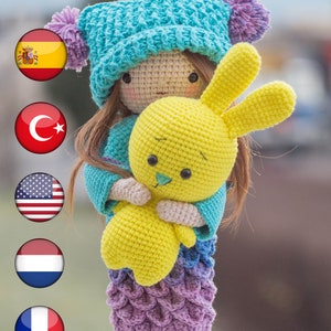 May include: A handmade crochet doll wearing a turquoise hat with purple pom-poms, holding a yellow crochet bunny. The doll has a turquoise sweater and a skirt that fades from purple to blue. The doll's hair is brown. The doll is a handmade item.