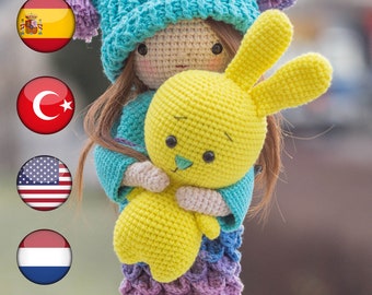 Puede incluir: Muñeca de ganchillo hecha a mano con un gorro turquesa con pompones morados, sosteniendo un conejito de ganchillo amarillo. La muñeca tiene un suéter turquesa y una falda que se desvanece de morado a azul. El pelo de la muñeca es castaño.