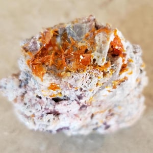May include: A close-up of a mineral specimen with a rough, porous texture. The rock is primarily white and gray, with vibrant orange crystalline formations throughout. The specimen has a layered appearance.