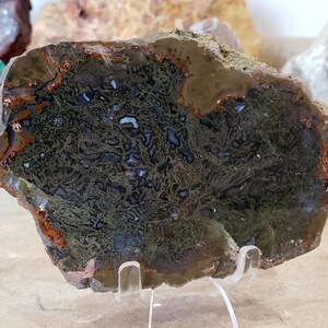 May include: A polished, irregularly shaped stone with a complex pattern of dark green, brown, and white inclusions. The stone is displayed on a clear acrylic stand, showcasing its natural textures and colors. Other stones are visible in the background.