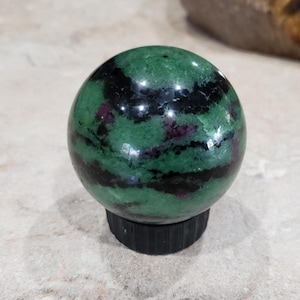 May include: A polished sphere of green zoisite with black tourmaline inclusions and flecks of ruby. The gemstone ball sits on a black, ridged base. The sphere is a decorative item.