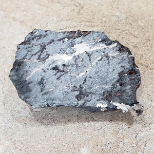 May include: A flat, irregularly shaped piece of dark gray metallic rock with a rough, textured surface. The stone features visible streaks of silver-toned mineral deposits and small pits across its face, with dark brown edges along the perimeter.