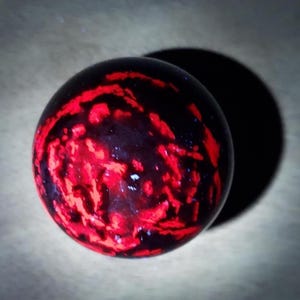 Puede incluir: Objeto esférico con fondo oscuro y llamativo patrón rojo. El diseño rojo se asemeja a venas o una red en la superficie. El objeto proyecta una sombra oscura.