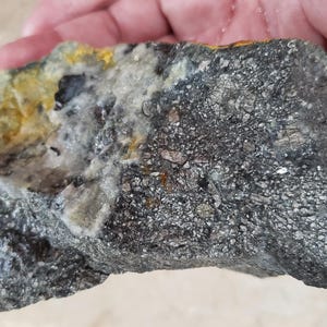 May include: A raw mineral specimen featuring a dark, metallic, and crystalline surface texture on one side, transitioning into a lighter, translucent, and yellowish-white quartz-like section with visible golden-colored mineral inclusions.