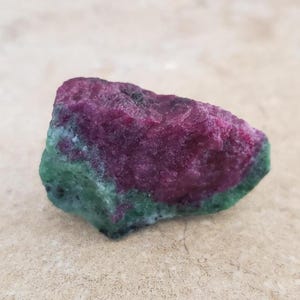 Puede incluir: Un espécimen mineral en bruto y sin pulir que presenta una sección de rubí rojo magenta intenso que contrasta con una matriz de zoisita verde vibrante. La piedra tiene una forma irregular y dentada con una textura cristalina rugosa.