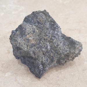 May include: A dark gray, rough-textured rock with a mix of light and dark areas. The rock has an irregular shape and appears to be a natural stone specimen. The surface has a coarse, uneven texture.