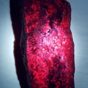 May include: A roughly shaped, reddish-purple rock with a textured surface. The stone has a vibrant, glowing appearance, with areas of deep red and dark purple. The overall shape is irregular, with a natural, unpolished look.