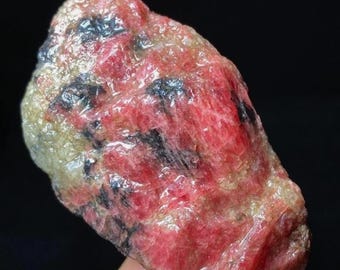 May include: A rough-cut gemstone with a mix of red, black, and tan colors. The stone has a natural, unpolished appearance, with visible textures and variations in color. The overall shape is irregular, with a slightly rounded form.