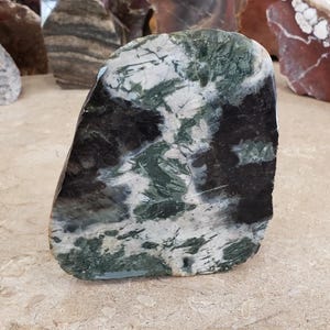 May include: A polished, irregularly shaped stone with a striking pattern of black, white, and green veining. The stone has a smooth, glossy surface and a natural, organic form. Other stones are visible in the background.