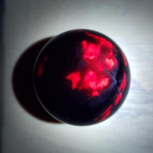May include: A dark, spherical object with a polished surface. The sphere has a deep black base color, with vibrant red and orange patterns across its surface. The patterns resemble glowing, irregular shapes. The object casts a dark shadow.
