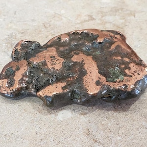 May include: A raw, irregular copper nugget with a metallic, reddish-brown surface. The object features a textured, uneven form with dark, oxidized crevices and smooth, polished copper highlights across its organic, molten-like shape.