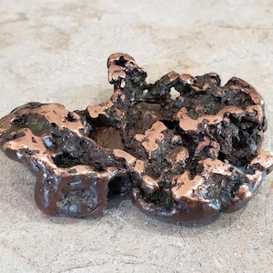 May include: A raw, irregular copper-colored metal nugget with a highly textured, porous surface. The object features a mix of shiny, polished metallic copper highlights and deep, dark, oxidized crevices throughout its organic, molten-like form.