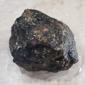Pode incluir: Uma rocha cinza escura com manchas douradas. A superfície áspera e texturizada da pedra é visível, com várias tonalidades de cinza e dourado. A rocha tem uma forma irregular.