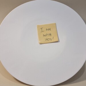 Può includere: Un grande piatto rotondo in ceramica bianca con un piccolo post-it quadrato giallo al centro. Il post-it riporta il messaggio scritto a mano "I AM WITH YOU!" in inchiostro nero. Il piatto è pulito e semplice.