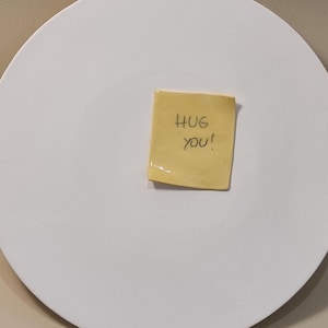 Può includere: Un grande piatto rotondo in ceramica bianca con un bordo leggermente irregolare. Una piccola nota adesiva gialla è attaccata al centro, con le parole scritte a mano "HUG YOU!" in inchiostro nero. Il piatto è posto su uno sfondo neutro.