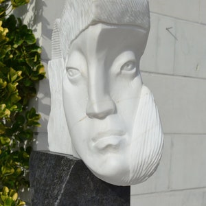 Puede incluir: Escultura de mármol blanco de la cabeza de una persona con rasgos faciales detallados. La escultura está sobre una base negra.