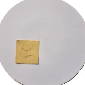 Può includere: Un grande piatto rotondo in ceramica bianca con un piccolo post-it quadrato giallo attaccato. Il biglietto ha un volto sorridente disegnato e le lettere "xoxo" sotto. Il piatto è semplice e liscio.
