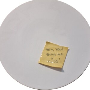 Può includere: Un piatto rotondo in ceramica bianca con un post-it giallo attaccato. Il biglietto dice "Hey, you! Give me a kiss!" in testo scritto a mano nero. Il piatto ha una superficie liscia.