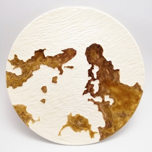 Può includere: Un piatto rotondo in ceramica bianco sporco con una superficie strutturata e disegni marroni astratti a forma di mappa. Il piatto presenta un motivo strutturato a forma di onda sulla superficie bianca, con forme marroni irregolari che assomigliano a continenti.