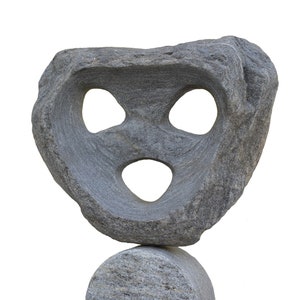 Puede incluir: Una escultura de piedra gris con tres agujeros tallados en ella. La escultura está equilibrada sobre una piedra redonda más pequeña.