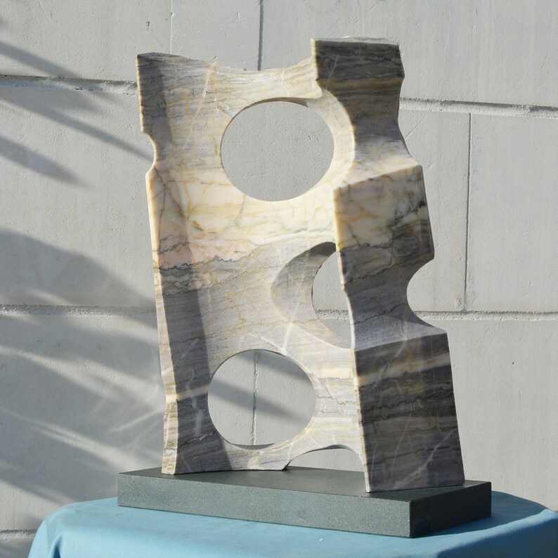 K&ouml;nnte beinhalten: Abstrakte Marmorskulptur mit drei kreisf&ouml;rmigen Ausschnitten. Der Stein weist eine Mischung aus grauen, beigen und wei&szlig;en Adern auf. Die Skulptur ist auf einem rechteckigen, dunkelgrauen Sockel montiert und wird vor einer wei&szlig;en Backsteinmauer ausgestellt.