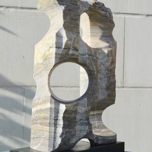 K&ouml;nnte beinhalten: Eine geschnitzte Steinskulptur mit einer kreisf&ouml;rmigen &Ouml;ffnung und einem rechteckigen Sockel. Die Skulptur besteht aus geschichtetem, mehrfarbigem Stein mit Graut&ouml;nen, Beige und Wei&szlig;. Der Sockel ist dunkelgr&uuml;n. Die Skulptur wird auf einer blauen Oberfl&auml;che pr&auml;sentiert.