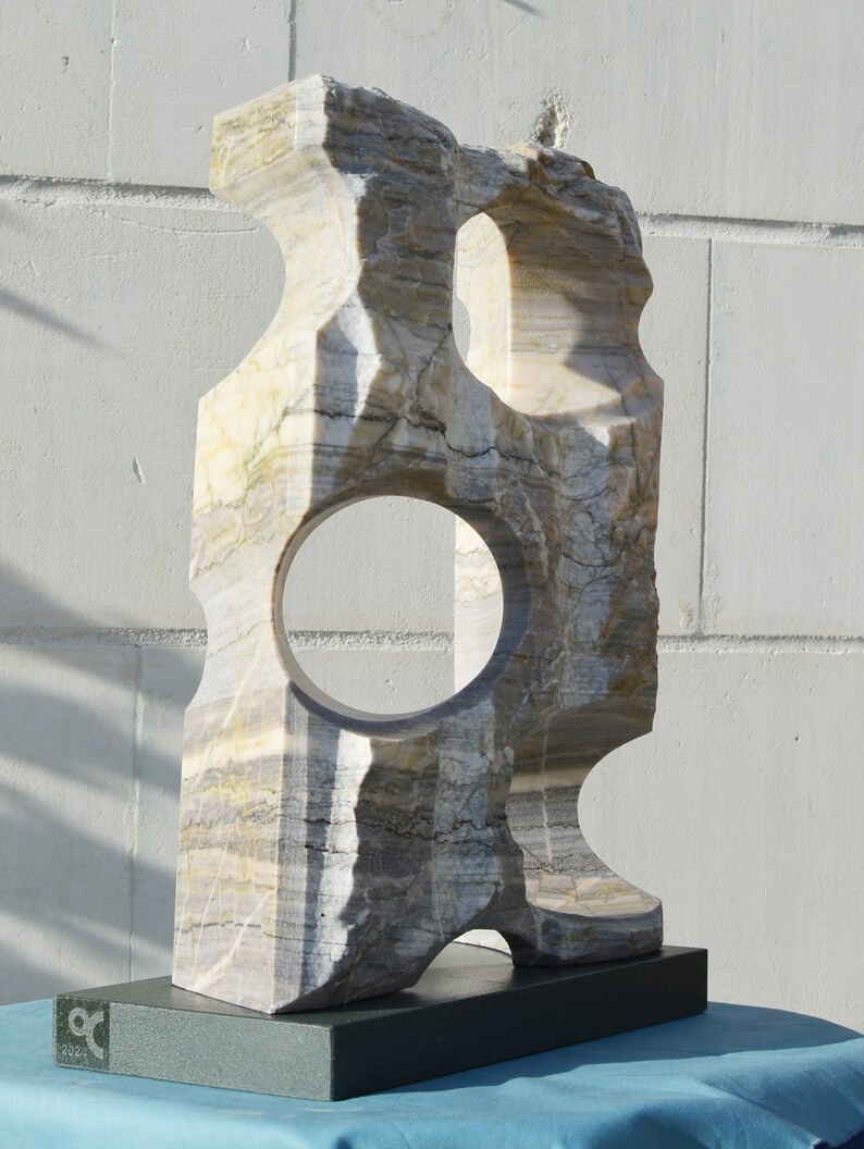 K&ouml;nnte beinhalten: Eine geschnitzte Steinskulptur mit einer kreisf&ouml;rmigen &Ouml;ffnung und einem rechteckigen Sockel. Die Skulptur besteht aus geschichtetem, mehrfarbigem Stein mit Graut&ouml;nen, Beige und Wei&szlig;. Der Sockel ist dunkelgr&uuml;n. Die Skulptur wird auf einer blauen Oberfl&auml;che pr&auml;sentiert.