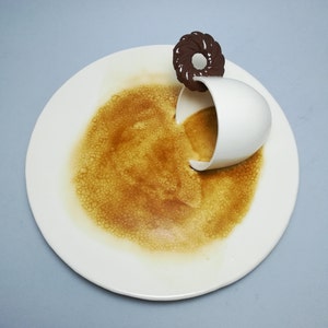 Può includere: Una tazza da caffè in ceramica bianca con un manico a forma di ciambella marrone è rovesciata su un piatto bianco, versando caffè marrone sul piatto.