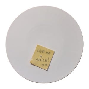 Può includere: Un piatto bianco e rotondo con un post-it giallo. Il post-it recita "GIVE ME A SMILE!" con un pennarello nero, con "xoxo" sotto. Il piatto è semplice e liscio.