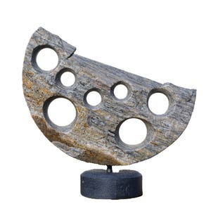Puede incluir: Escultura abstracta de piedra con forma semicircular. La piedra gris y marrón tiene varios agujeros circulares. Está montada sobre una base negra. Una pieza decorativa.