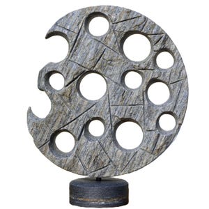 Puede incluir: Una escultura de piedra gris con forma circular y numerosas aberturas circulares. La escultura tiene una superficie texturizada y está montada sobre una base cilíndrica gris oscuro. El diseño presenta una serie de líneas que se cruzan.