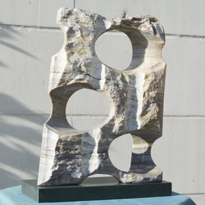 K&ouml;nnte beinhalten: Abstrakte Steinskulptur mit drei kreisf&ouml;rmigen Ausschnitten. Die Skulptur besteht aus wei&szlig;em, grauem und beigem Marmor und steht auf einem dunkelgr&uuml;nen rechteckigen Sockel. Die Skulptur hat eine raue, strukturierte Oberfl&auml;che und wird im Freien ausgestellt.