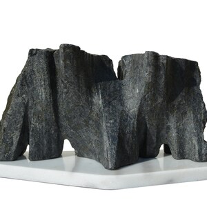 Puede incluir: Escultura abstracta hecha de dos piezas de piedra gris oscuro, montada sobre una base de mármol blanco.