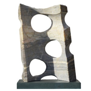 Puede incluir: Escultura abstracta de piedra con tres cortes ovalados. La escultura está hecha de mármol gris, beige y blanco y está montada sobre una base rectangular gris oscuro. La piedra tiene vetas naturales y una superficie texturizada.