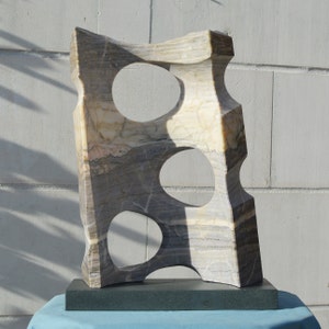 K&ouml;nnte beinhalten: Abstrakte Steinskulptur mit drei kreisf&ouml;rmigen Ausschnitten. Die Skulptur besteht aus geschichtetem beigem, grauem und braunem Stein, montiert auf einem dunkelgrauen rechteckigen Sockel. Das St&uuml;ck hat eine moderne, k&uuml;nstlerische &Auml;sthetik.