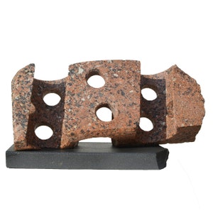 May include: Abstract sculpture made of reddish-brown granite with black speckles. The sculpture has a series of circular holes and is mounted on a black rectangular base. The stone has a rough, textured surface.