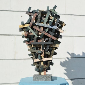 Puede incluir: Escultura abstracta hecha de piezas de granito apiladas e interconectadas en tonos de negro, marrón, verde y beige. La escultura está montada sobre una base cuadrada de granito.