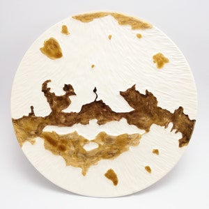 Può includere: Un piatto rotondo in ceramica bianco sporco con superficie strutturata. Presenta un design astratto marrone e beige che ricorda un paesaggio. Il design include aree in rilievo e alcune piccole forme sparse. Un oggetto decorativo.