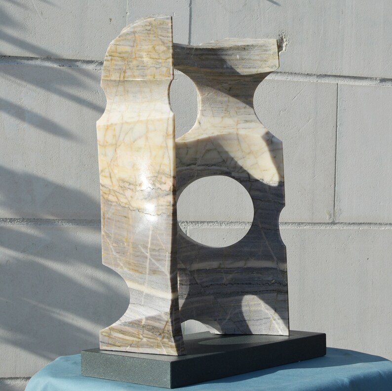 K&ouml;nnte beinhalten: Abstrakte Marmorskulptur in Hellbeige und Grau. Die rechteckige Form weist zwei kreisf&ouml;rmige Ausschnitte auf und steht auf einem dunkelgrauen Sockel. Der Stein weist nat&uuml;rliche Maserungen auf. Die Skulptur wird im Freien ausgestellt.