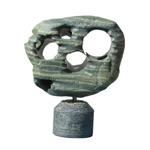 Puede incluir: Escultura abstracta de piedra gris-verdosa con múltiples aberturas circulares. La escultura se apoya sobre una base cilíndrica de piedra. La piedra presenta un aspecto texturizado y estratificado, creando una estética natural y orgánica.