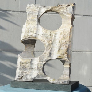 K&ouml;nnte beinhalten: Eine geschnitzte Steinskulptur mit drei kreisf&ouml;rmigen &Ouml;ffnungen. Die Skulptur ist rechteckig mit einer rauen, strukturierten Oberfl&auml;che in Beige-, Grau- und Wei&szlig;t&ouml;nen. Sie ist auf einem dunkelgrauen rechteckigen Sockel montiert. Die Skulptur wird im Freien ausgestellt.