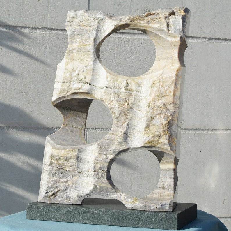 K&ouml;nnte beinhalten: Eine geschnitzte Steinskulptur mit drei kreisf&ouml;rmigen &Ouml;ffnungen. Die Skulptur ist rechteckig mit einer rauen, strukturierten Oberfl&auml;che in Beige-, Grau- und Wei&szlig;t&ouml;nen. Sie ist auf einem dunkelgrauen rechteckigen Sockel montiert. Die Skulptur wird im Freien ausgestellt.