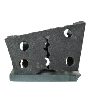 Pode incluir: Uma escultura de granito preto com um design irregular e partido. A escultura tem vários orifícios circulares e é exibida numa base mais pequena de cor cinzento escuro.