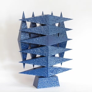 Könnte beinhalten: Blaue geometrische Skulptur mit strukturierter Oberfläche. Sie zeigt gestapelte Diamantformen und dreieckige Spitzen. Die Skulptur ist auf einem quadratischen Sockel montiert. Das Design ist abstrakt und modern.