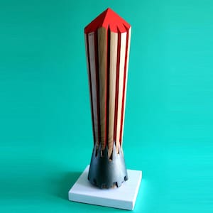 Puede incluir: Un objeto decorativo de madera con una parte superior geométrica roja y una base negra, colocado sobre un cuadrado blanco. El cuerpo presenta rayas verticales alternas de madera natural y rojo. El fondo es de color turquesa sólido.