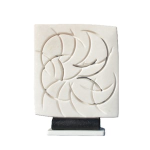Puede incluir: Una escultura cuadrada blanca con un diseño abstracto tallado de formas curvas superpuestas. La escultura se asienta sobre una base rectangular negra sobre una base cuadrada blanca. La obra de arte es un diseño moderno y minimalista.