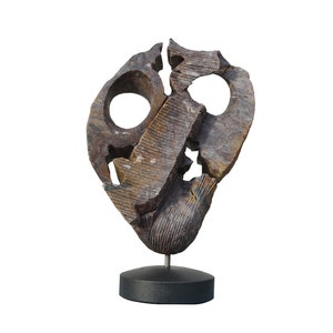 Puede incluir: Escultura abstracta hecha de piedra marrón y gris con una superficie rugosa y texturizada. La escultura tiene dos agujeros grandes y está montada sobre un pedestal negro.