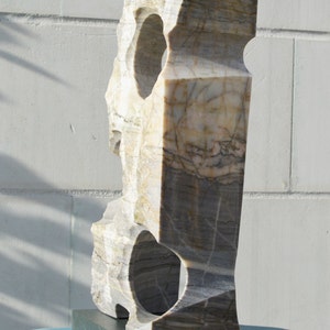 K&ouml;nnte beinhalten: Eine geschnitzte Steinskulptur mit rechteckiger Form und zwei kreisf&ouml;rmigen &Ouml;ffnungen. Die Skulptur besteht aus einem hellen Stein mit grauen und braunen Adern und steht auf einem dunkelgrauen rechteckigen Sockel.