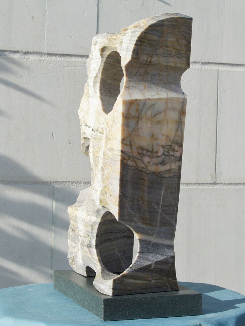 K&ouml;nnte beinhalten: Eine geschnitzte Steinskulptur mit rechteckiger Form und zwei kreisf&ouml;rmigen &Ouml;ffnungen. Die Skulptur besteht aus einem hellen Stein mit grauen und braunen Adern und steht auf einem dunkelgrauen rechteckigen Sockel.